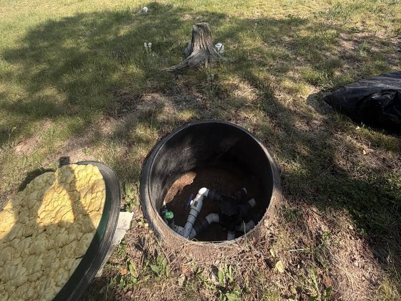 Open septic tank access in a grassy yard, with a pond and pine trees in the background